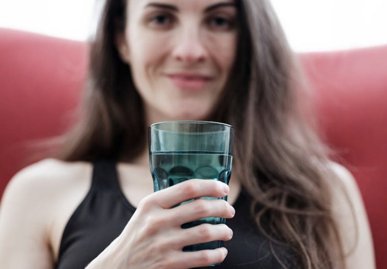 Smiling woman with long hair holding a glass of water, seated indoors with a red chair.